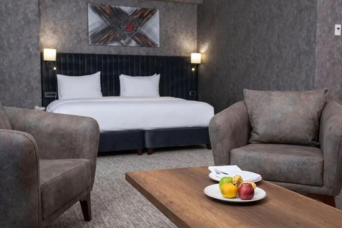 A hotel room with a bed, two armchairs, a wooden coffee table with fruit, and a painting on the wall.