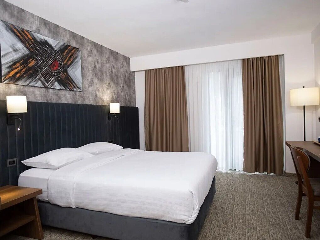 A hotel room with a bed, two bedside lamps, a desk, a chair, a large abstract painting, and a window with curtains.