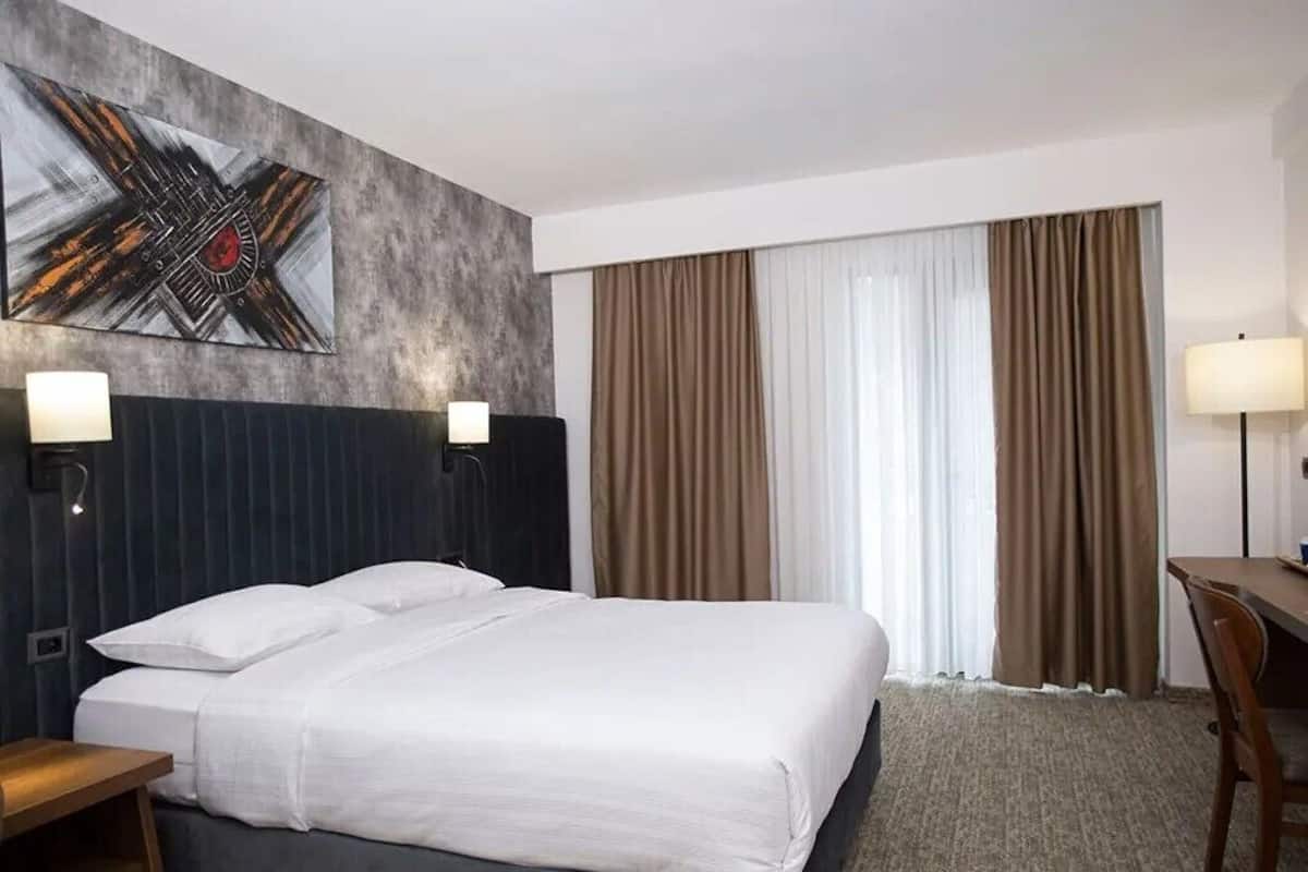 A hotel room with a bed, two bedside lamps, a desk, a chair, a large abstract painting, and a window with curtains.