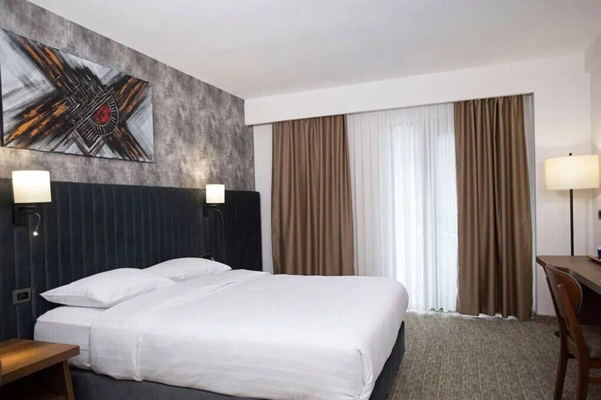 A hotel room with a bed, two bedside lamps, a desk, a chair, a large abstract painting, and a window with curtains.