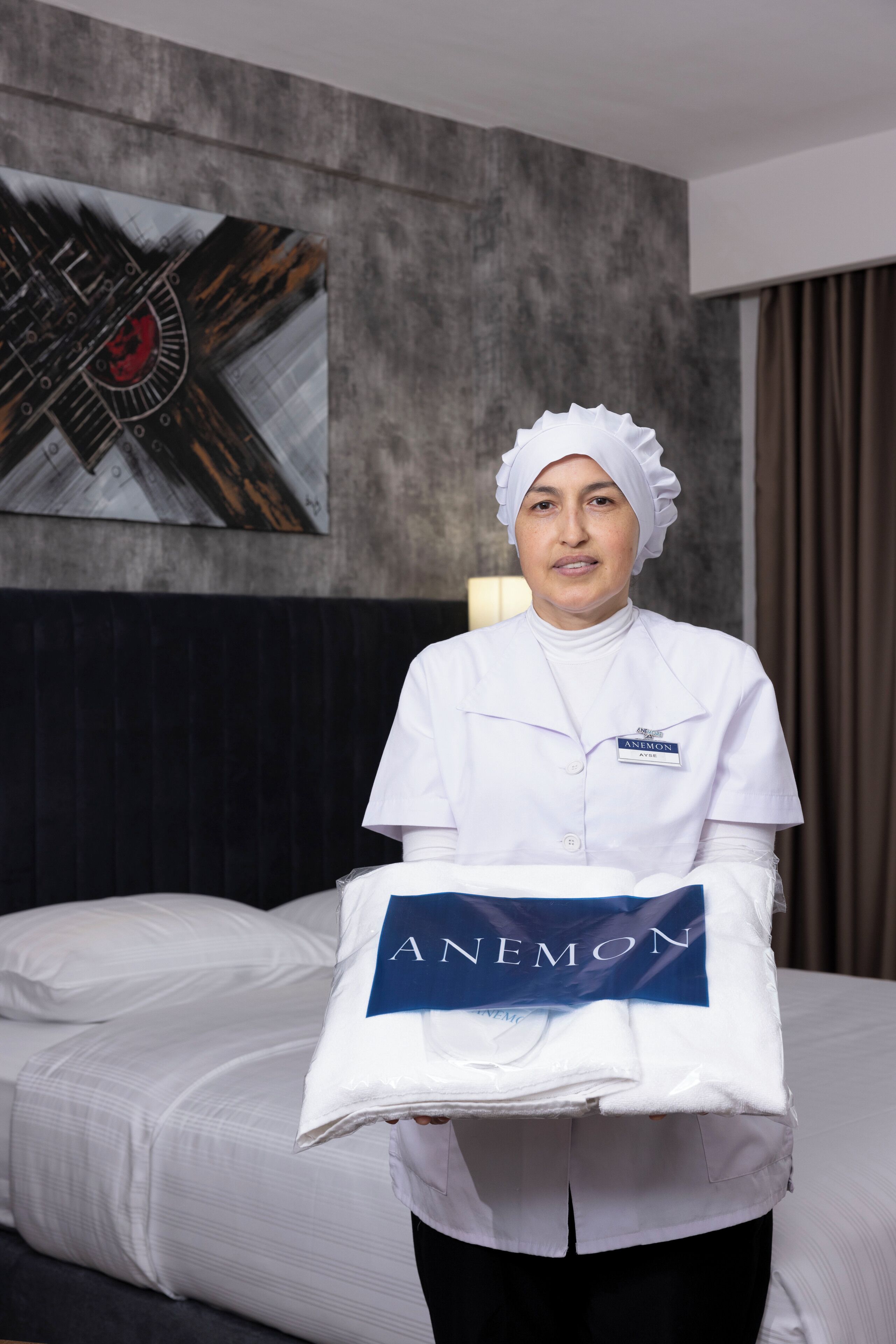 A person in a white uniform holding a towel with "ANEMON" printed on it, standing in a hotel room with a bed, a dark headboard, and a wall art piece.