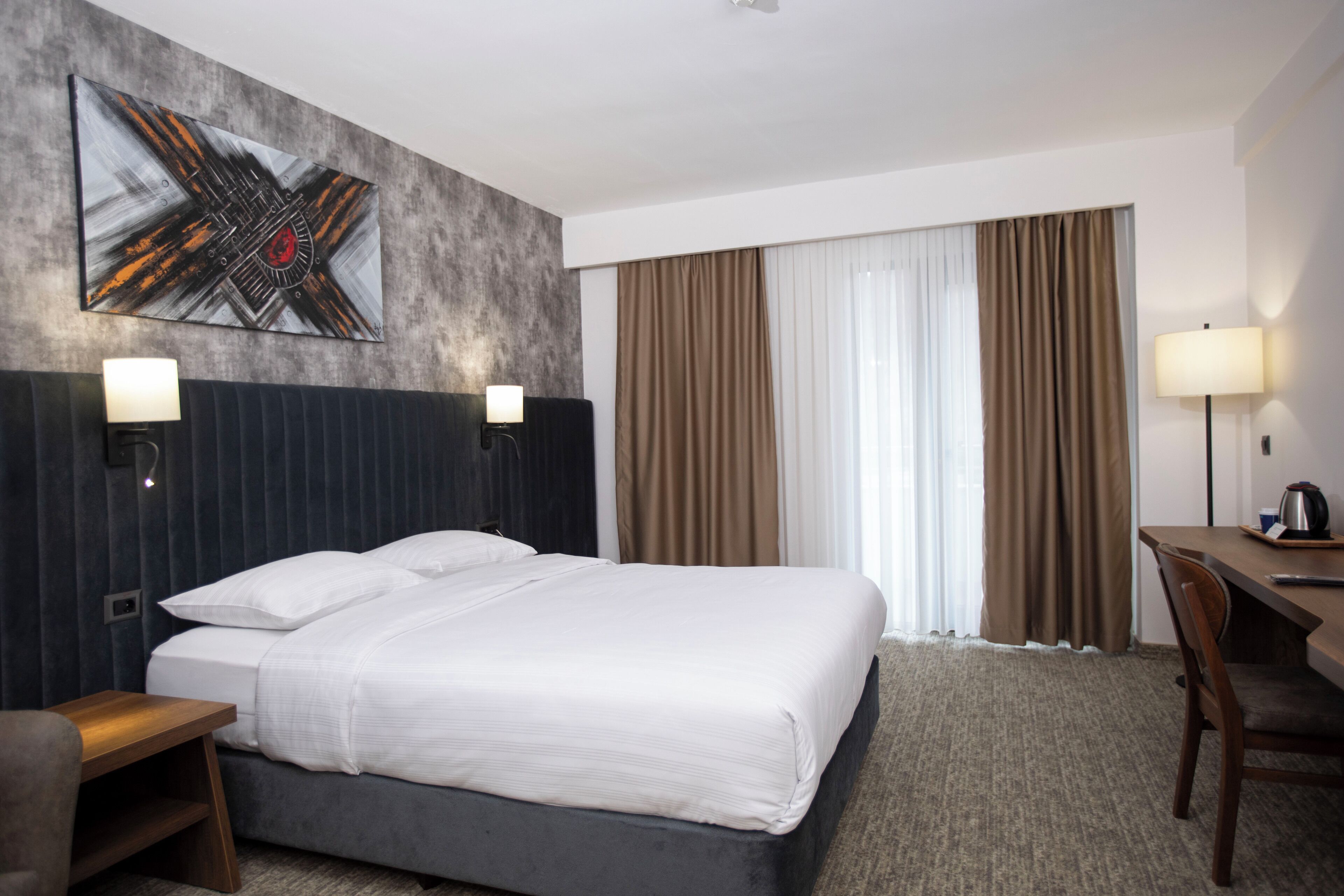 A hotel room with a large bed, two bedside tables, a desk, and a chair. There are two wall-mounted lamps, a painting on the wall, and a window with curtains.