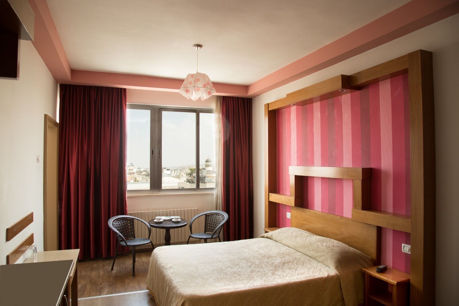 A hotel room with a bed, a desk, two chairs, a window with curtains, and a city view.