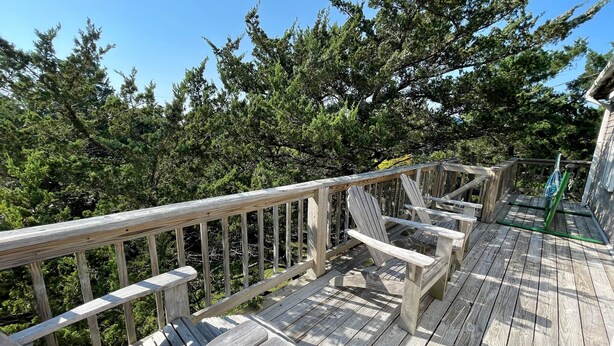 A wooden deck with Adirondack chairs and a glass table, surrounded by dense green trees. - Neaptide: Secluded & Spacious Home in Jackson Dunes (Ocracoke)