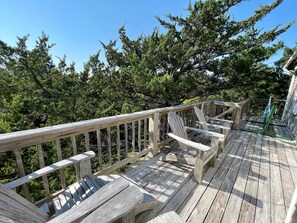 Una terraza de madera con sillas Adirondack y una mesa de vidrio, rodeada de árboles verdes y frondosos.
