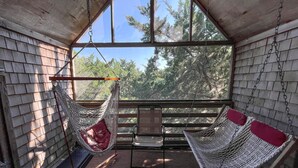 A hammock chair suspended from the ceiling in a wooden cabin with a view of trees through large windows. - Neaptide: Secluded & Spacious Home in Jackson Dunes (Ocracoke)
