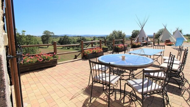 A patio with round tables and chairs, a wooden fence, and teepees in the background. - LES CHAMBRES DE L'OUEST (Longessaigne)