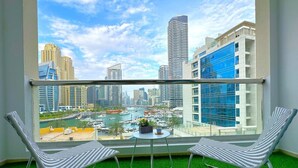 A balcony with two striped chairs, a small round table, and a view of a cityscape with tall buildings and a marina. - Big Studio w/ Balcony and easy access to the Beach in Marina (Dubai)