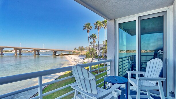A balcony with two white Adirondack chairs and a small table, overlooking a river with a bridge and palm trees.
