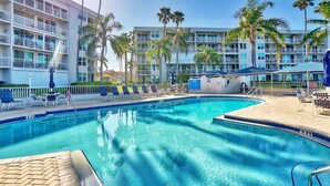 A swimming pool with lounge chairs and umbrellas, surrounded by a multi-story building with balconies and palm trees.