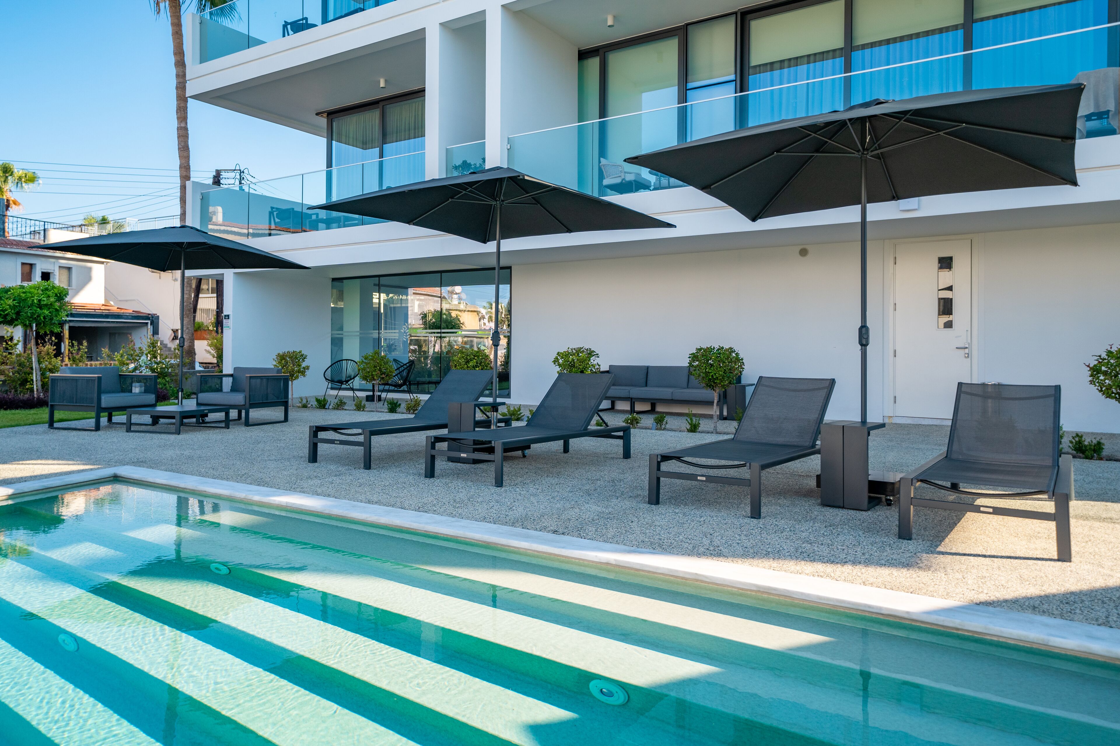Uma área moderna com piscina ao ar livre, cadeiras de sol e guarda-sóis, rodeada por um edifício de vários andares com amplas janelas e uma varanda.