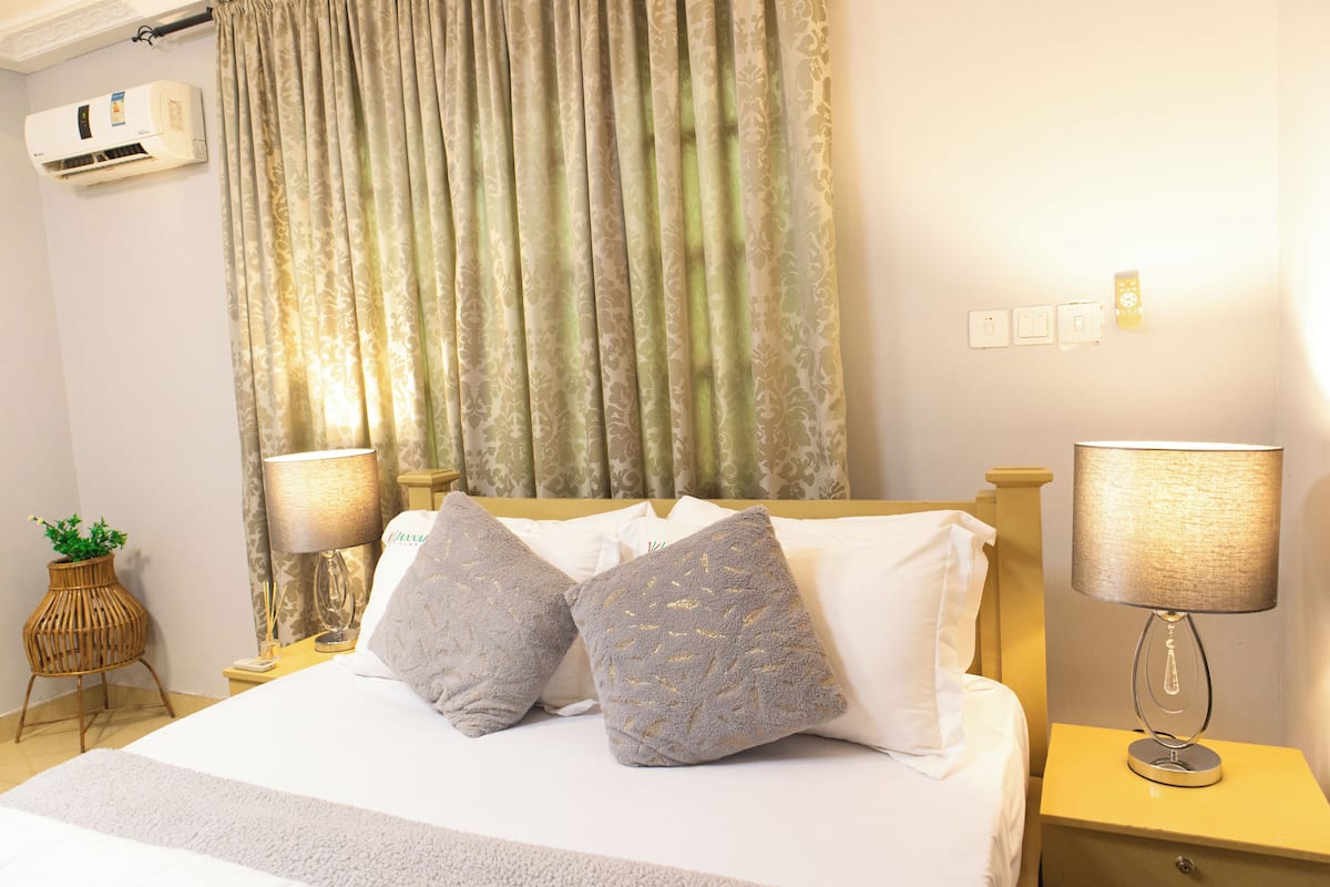 Cama bem arrumada com duas almofadas, luminária de cabeceira e ar-condicionado na parede.
