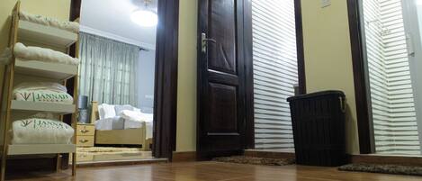 Une chambre d’hôtel avec un plancher en bois, un lit avec du linge de maison blanc, un banc en bois avec des serviettes, une porte et une poubelle.