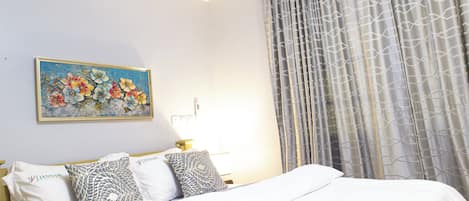 Una cama bien hecha con sábanas blancas, un cuadro floral en la pared y una cortina estampada.
