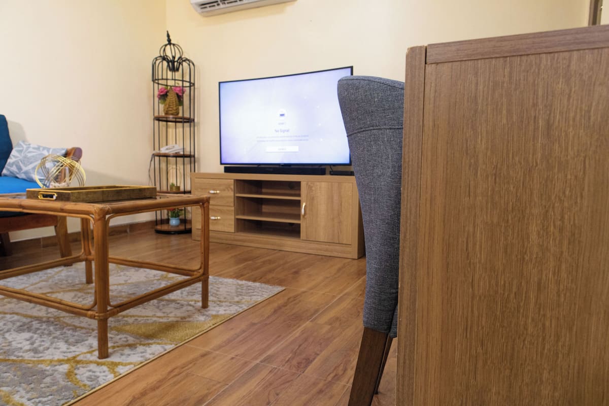 Uma sala de estar com piso de madeira, uma televisão em um móvel, um sofá cinza e uma pequena mesa com um cesto.