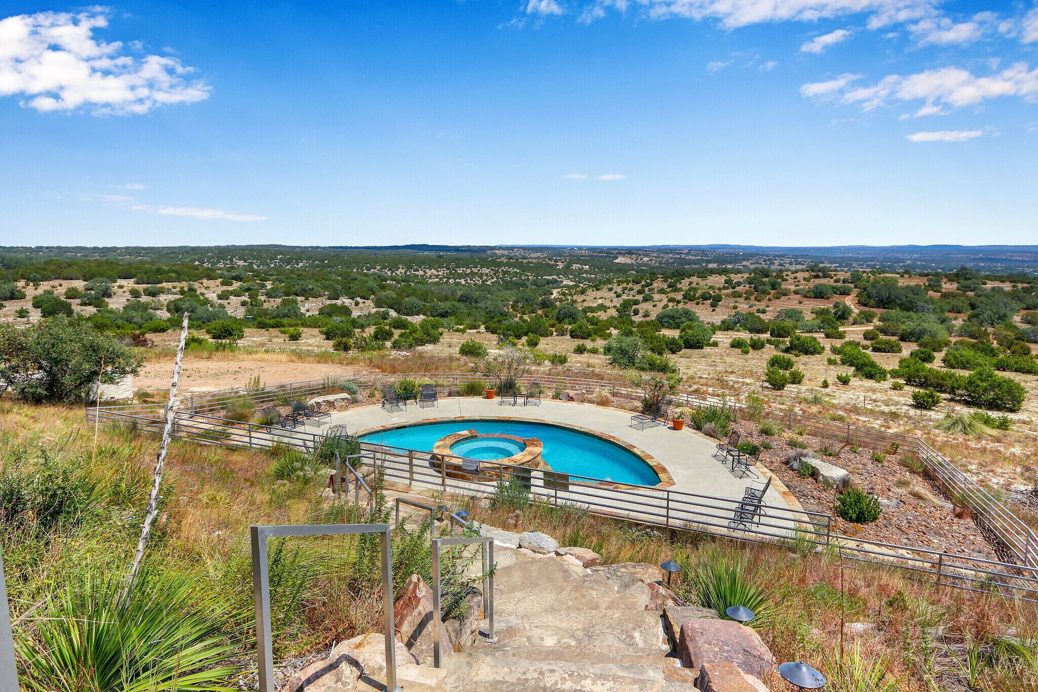 A swimming pool with a circular shape, surrounded by a metal railing, set in a dry, hilly landscape with sparse vegetation.
