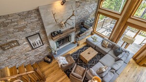 A spacious living area with a stone fireplace, a large window, and a wooden staircase. - Luxury 4BR Beehive Basin Home | Hot Tub, Mountain Views & Sleeps 13 (Big Sky)