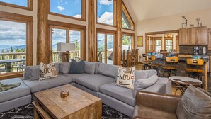 A spacious living area with a large sectional sofa, a wooden coffee table, and a view of mountains through large windows. - Luxury 4BR Beehive Basin Home | Hot Tub, Mountain Views & Sleeps 13 (Big Sky)