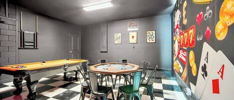 A pool table with chairs, a checkered floor, and a wall with playing card designs.