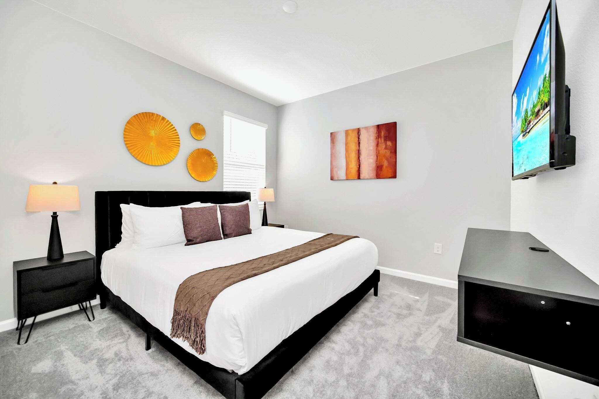 A modern bedroom with a large bed, bedside tables with lamps, a flat-screen TV mounted on the wall, and decorative wall art.