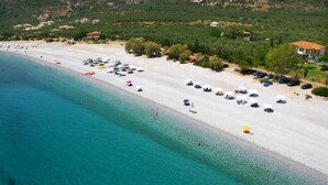 An aerial view of a beach with a row of beach umbrellas and sun loungers, a clear blue sea, and buildings in the background. - Eleonas Houses - Kardamili's  Bianca Poolside Gem (Kardamyli)