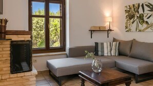 A living room with a fireplace, a sofa, a coffee table, and a window with a view of trees. - Eleonas Houses - Kardamili's  Bianca Poolside Gem (Kardamyli)