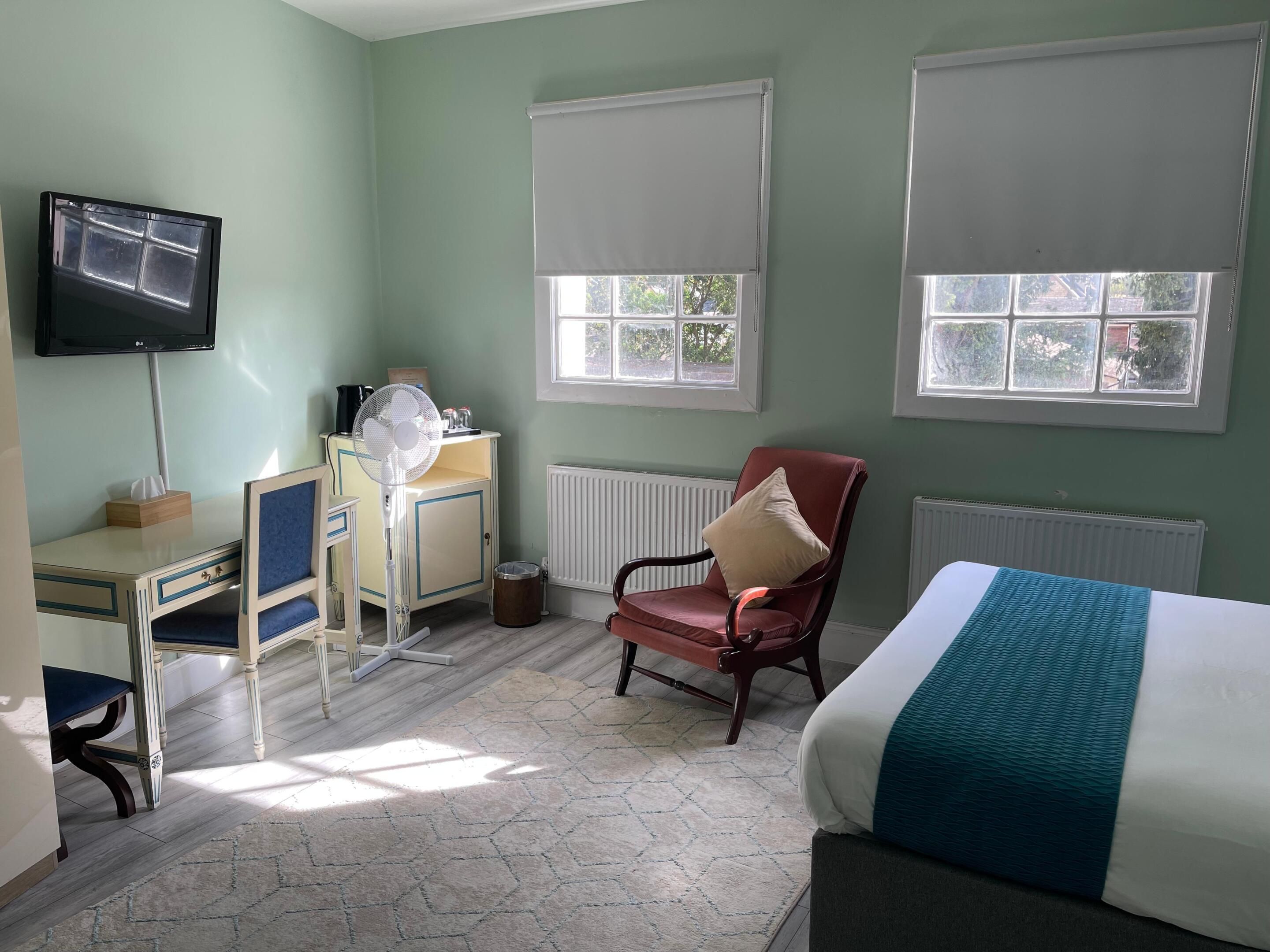 A hotel room with a bed, a desk with a computer, a chair, and two windows with blinds.
