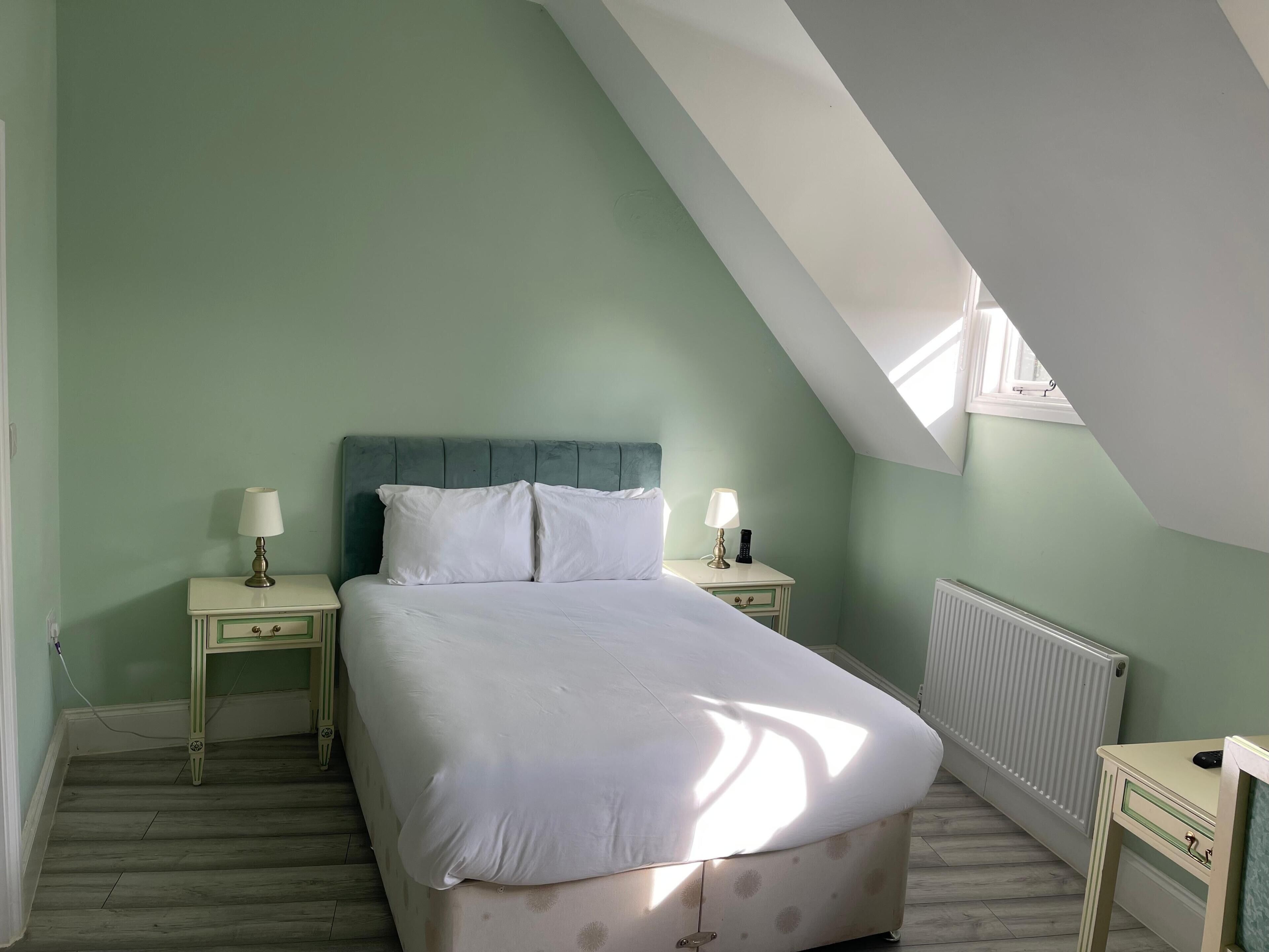 A bedroom with a sloped ceiling, a bed with white bedding, two bedside tables with lamps, and a radiator.