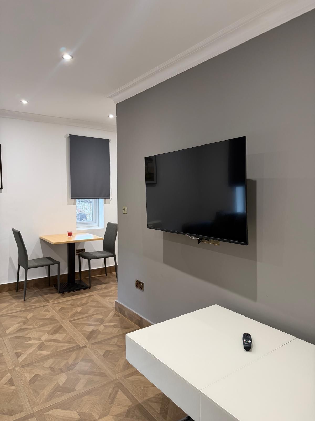 Una habitación moderna con una televisión de pantalla plana en la pared, un banco blanco, dos sillas, una mesa pequeña con una taza roja y un suelo de madera con diseño.