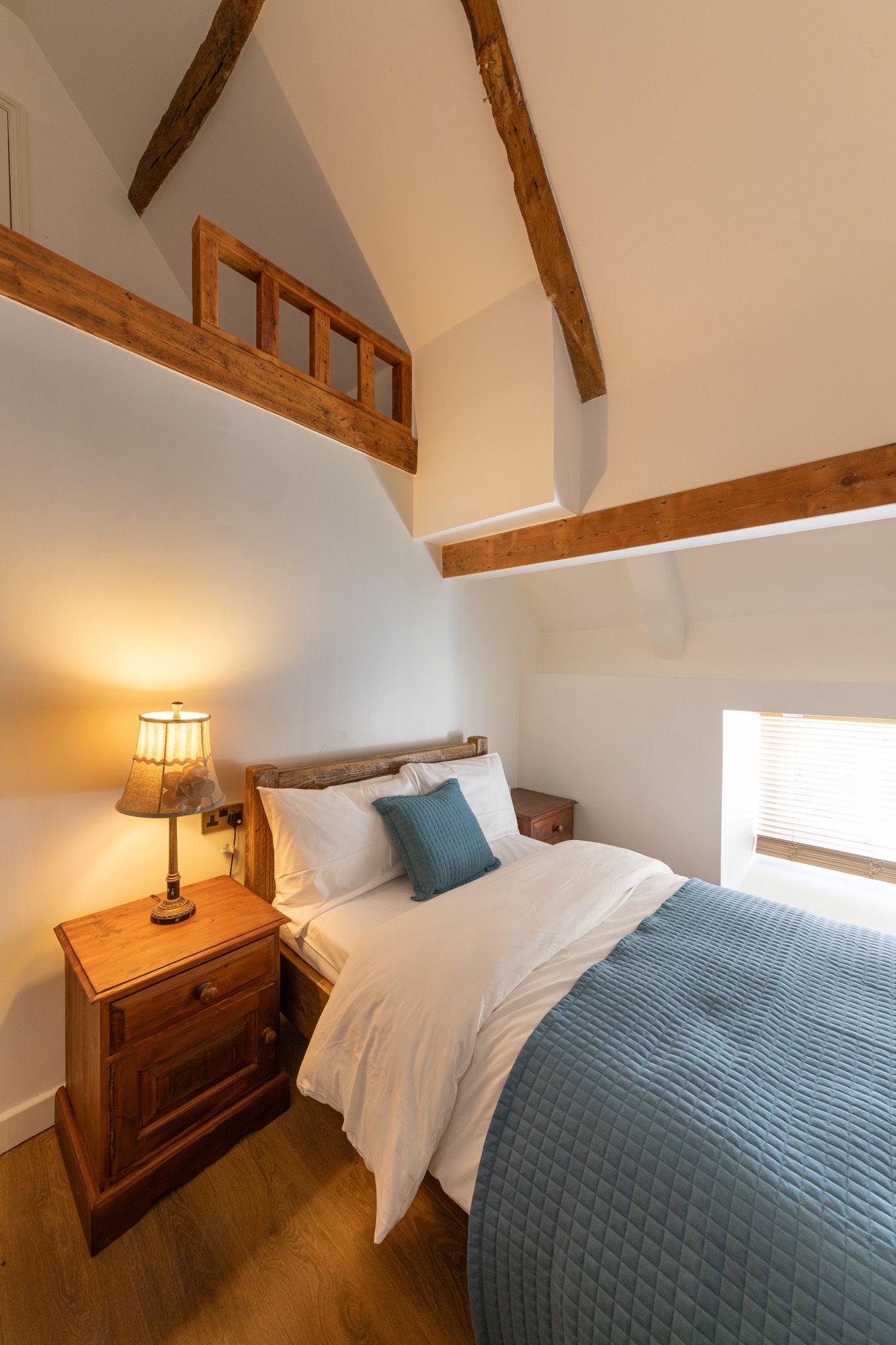 A bedroom with a wooden headboard, a bedside table with a lamp, a bed with white bedding and a blue quilt, and a window with blinds.