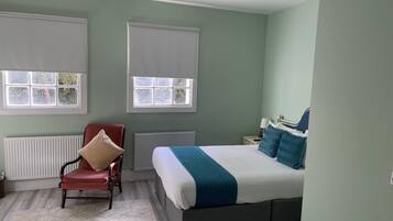 Quarto com cama, uma cadeira, duas janelas com persianas, um radiador e uma luminária no teto.