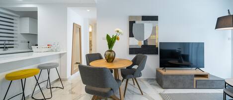 A modern living room with a round wooden table, grey upholstered chairs, a flat-screen TV, and a decorative vase with flowers.