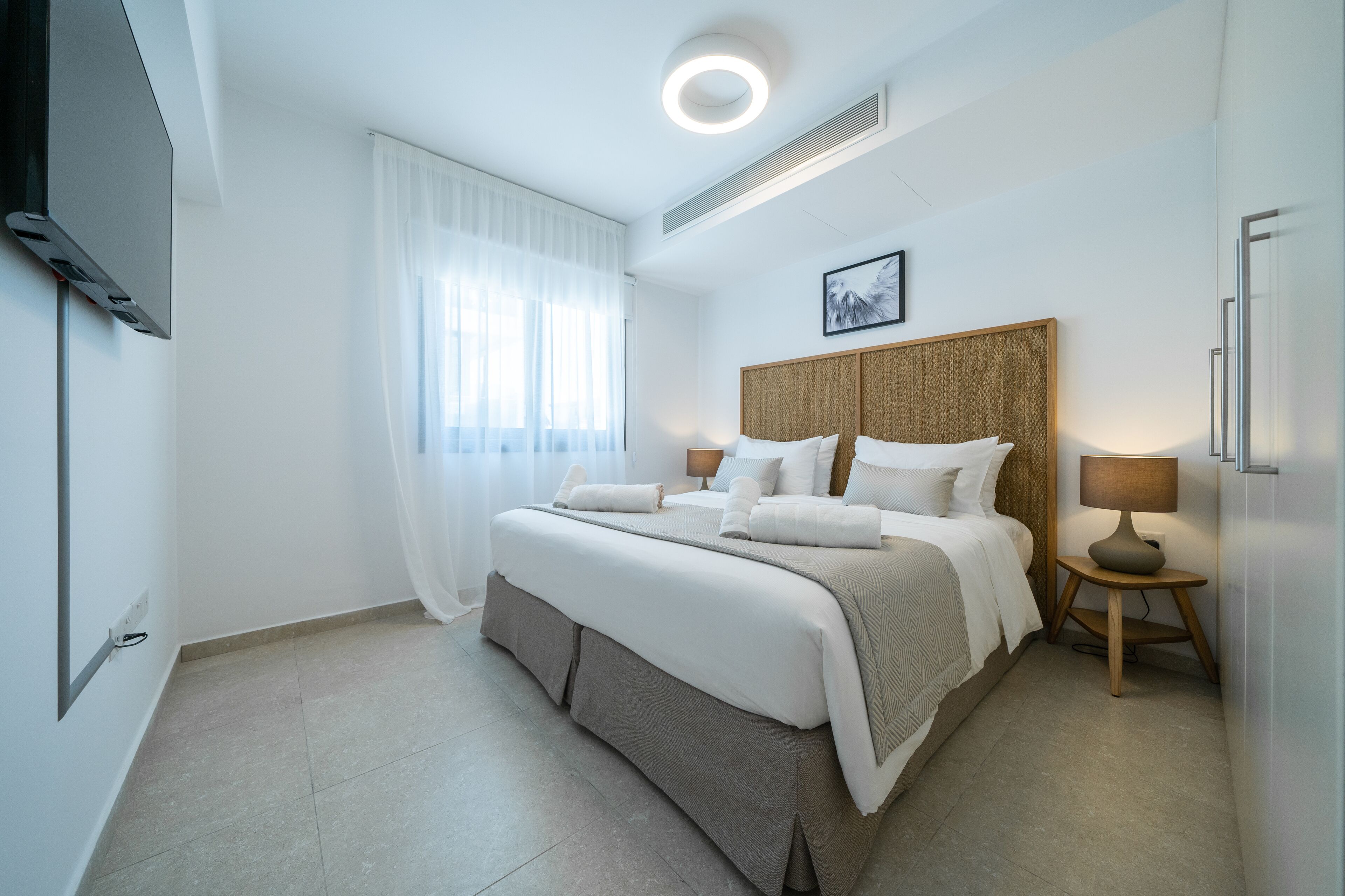 A modern hotel room with a large bed, a bedside table with a lamp, and a flat-screen TV mounted on the wall.