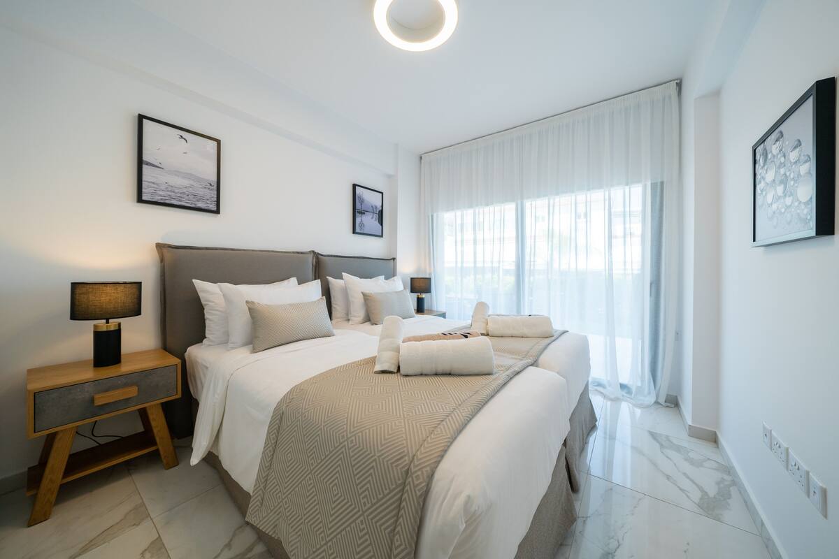 Une chambre d’hôtel moderne avec un grand lit, une table de chevet avec une lampe, une commode et une vue sur la mer par la fenêtre.