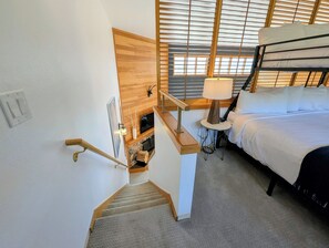 A bedroom with a bed, a nightstand, a lamp, a window with blinds, and a staircase leading to another room.