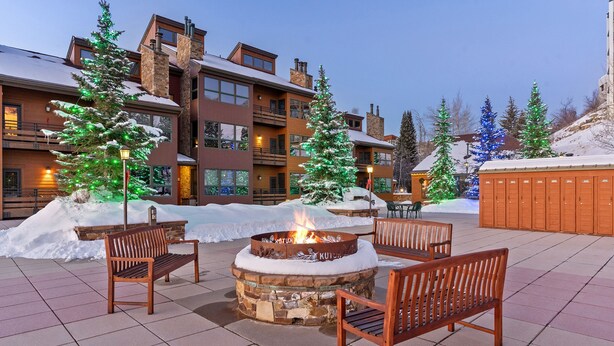 A fire pit with benches, surrounded by snow and trees with lights, in a courtyard with a building in the background.
