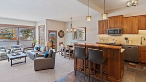 A spacious living area with a sofa, coffee table, and bar stools. A kitchen with wooden cabinets and a microwave is visible.