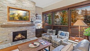 A cozy living room with a stone fireplace, a large framed landscape painting, and a view of snowy trees through large windows.