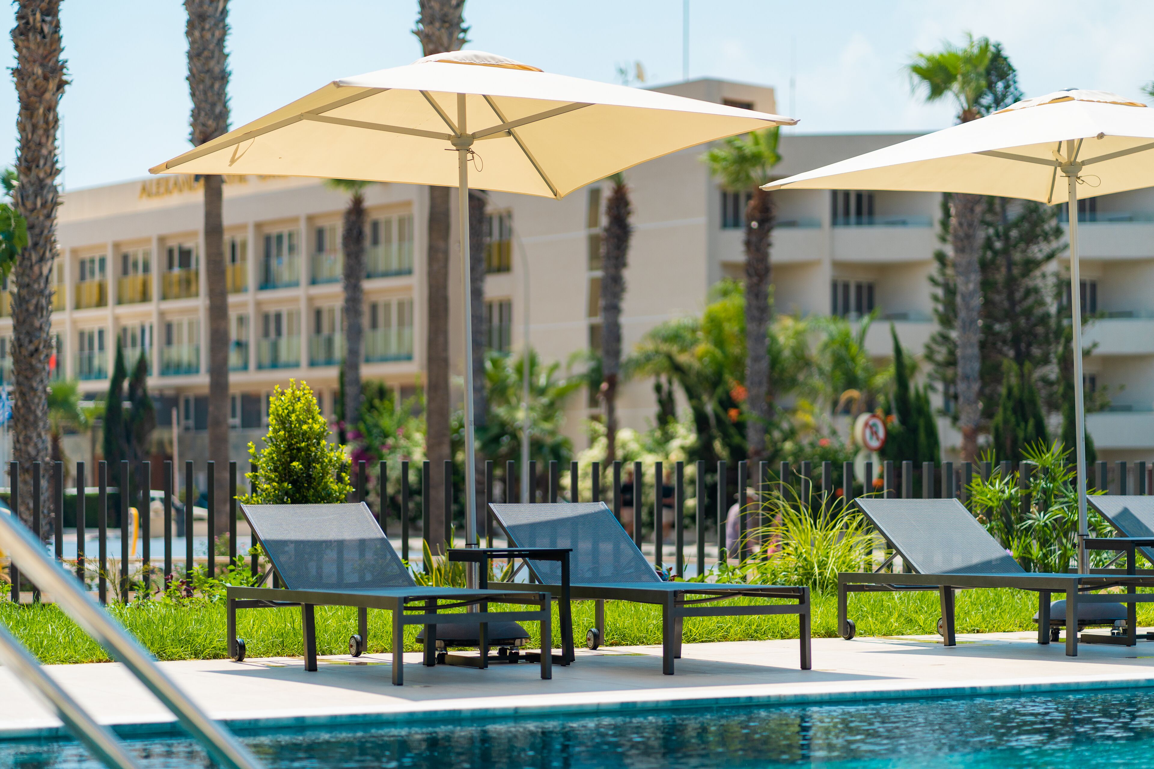 A poolside area with lounge chairs and umbrellas, a pool, and a building with multiple balconies in the background.