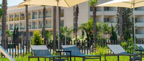 A poolside area with lounge chairs and umbrellas, a pool, and a building with multiple balconies in the background.