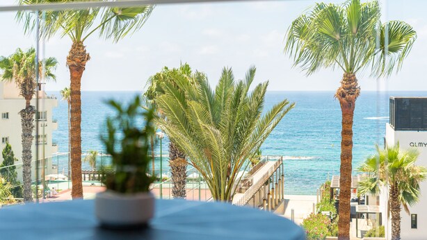 A balcony with a view of the sea, palm trees, and a building with the sign "ALMY". - Waterside by Deserve (Paphos)