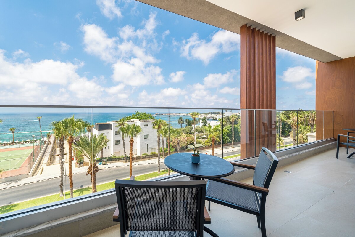 A balcony with a table and chairs, overlooking a coastal area with palm trees and buildings.
