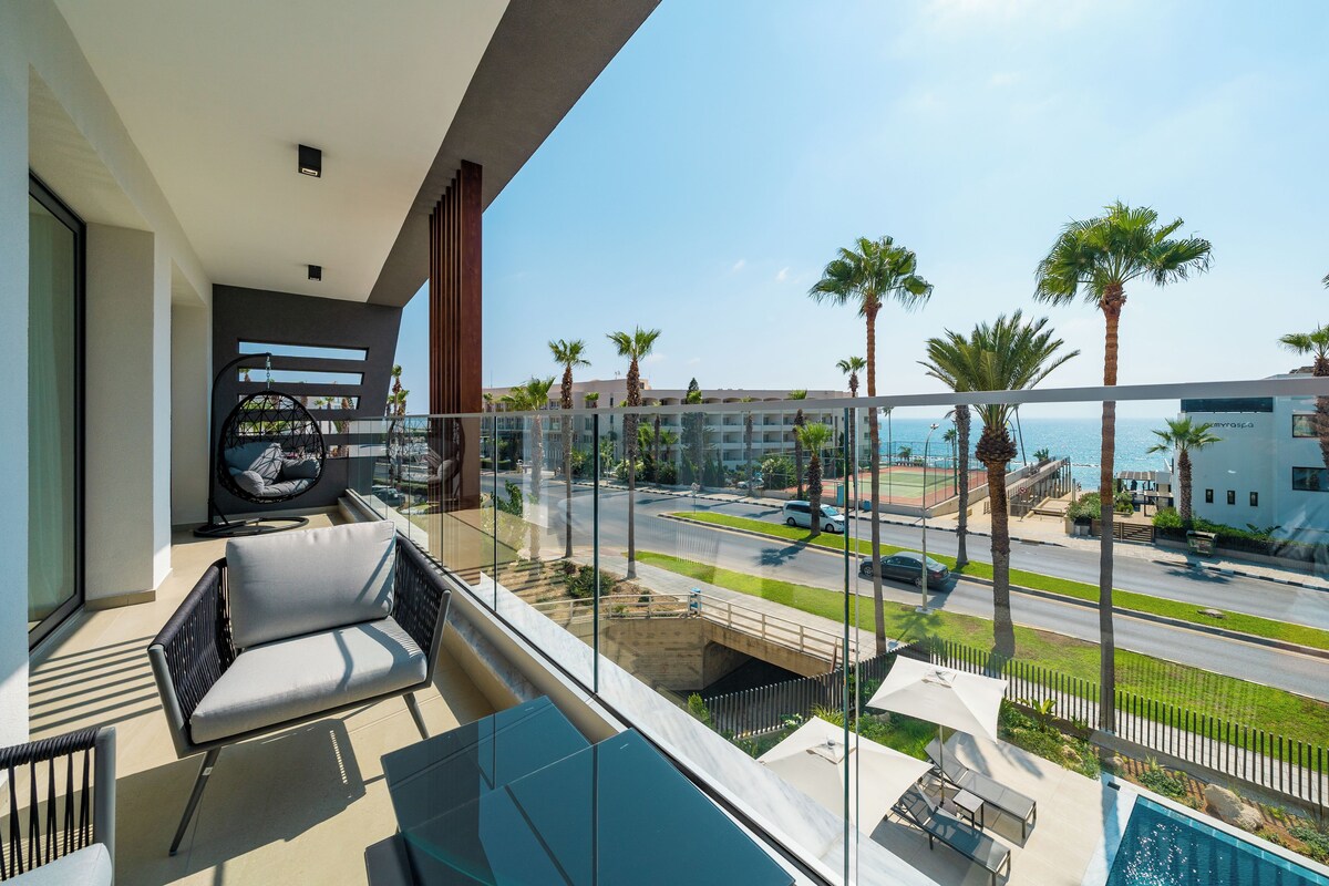 A balcony with a view of a road, palm trees, and a swimming pool.
