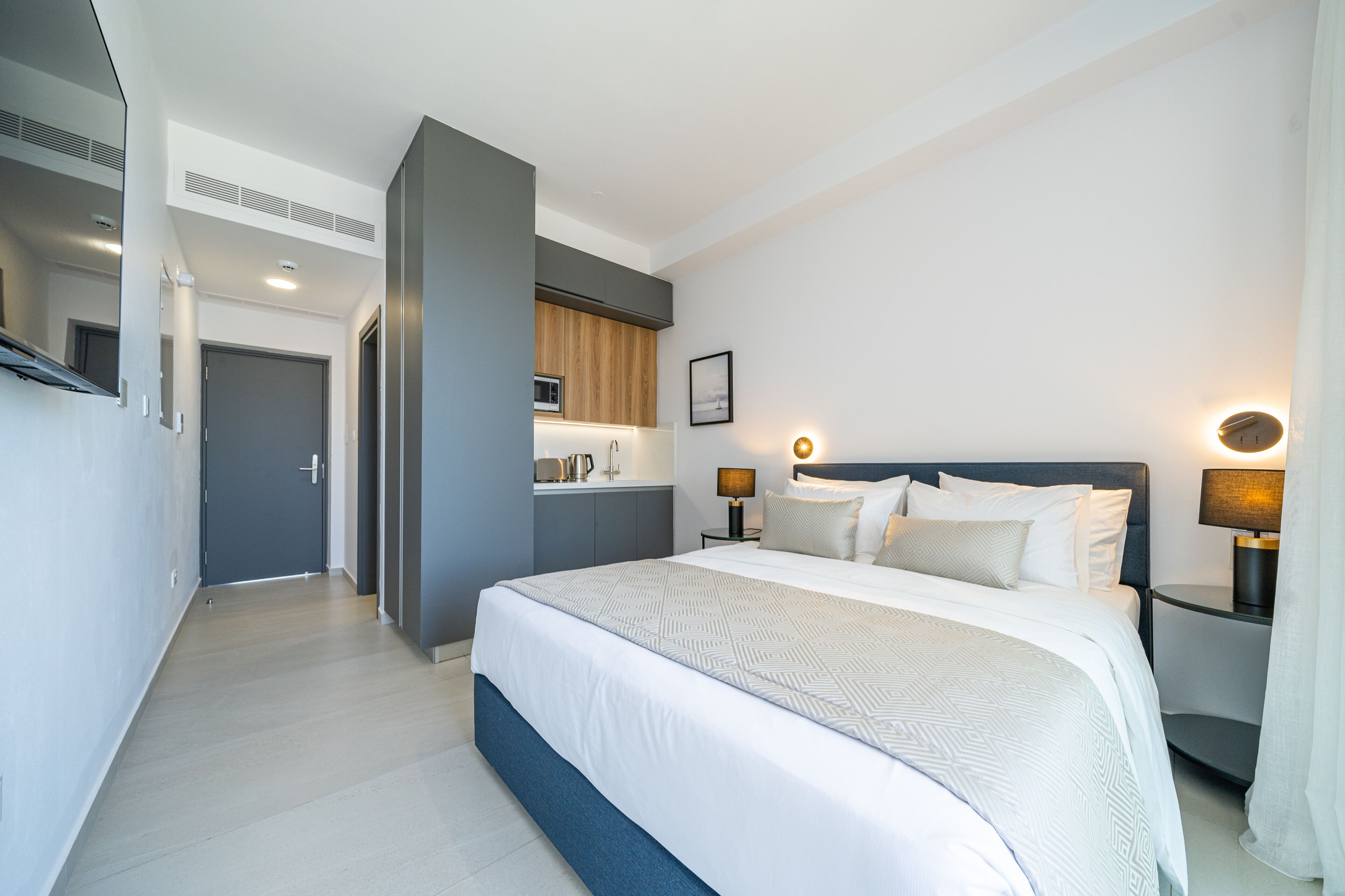 A modern hotel room with a large bed, bedside tables, a built-in kitchen, and a separate bathroom.