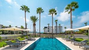 A swimming pool with lounge chairs and umbrellas, surrounded by palm trees and a building in the background. - Waterside by Deserve (Paphos)