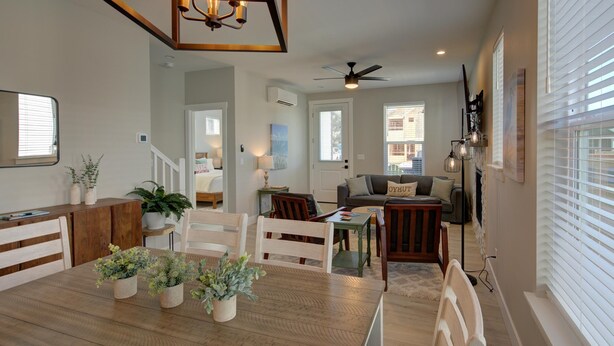A spacious living area with a wooden dining table, chairs, and potted plants. There is a ceiling fan, a sofa, and a TV mounted on the wall.