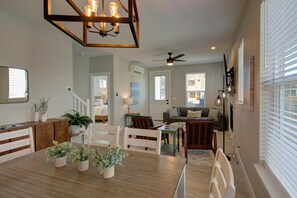 Un espacio habitable amplio con una mesa de comedor de madera, sillas y plantas en macetas. Hay un ventilador de techo, un sofá y un televisor montado en la pared.