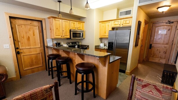 A kitchen with wooden cabinets, a stainless steel refrigerator, a microwave, and a stove. There are three stools and a table with chairs. - Silverado Lodge - 1 BR Suite, King Bed & Pool View by RedAwning (Park City)