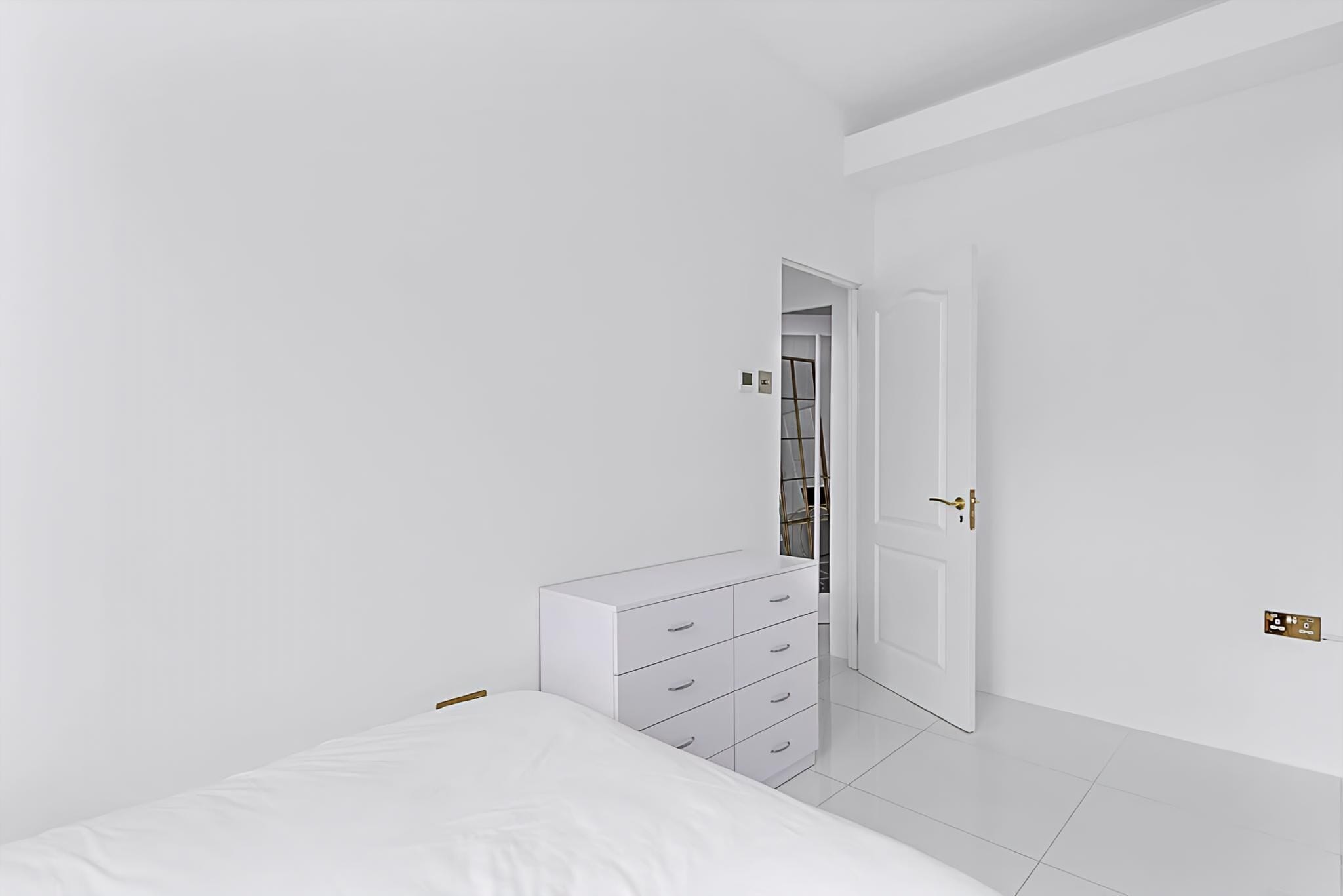 Une chambre à coucher au style minimaliste, avec un lit blanc, une commode à tiroirs et une porte donnant sur un placard.