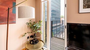 Ein modernes Wohnzimmer mit großem Fernseher, Stehlampe, Pflanze und Blick auf die Stadt durch eine Glastür.